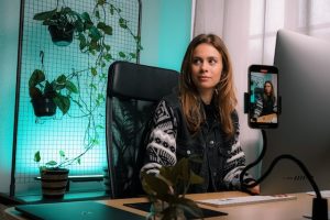 content creator- a women dressed in a black and white zipper jacket, sitting on a rolling chair and making a video with her phones back camera and her phone on a tripod