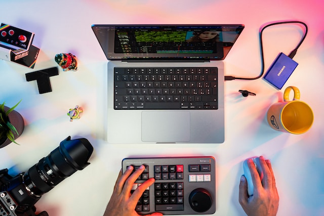 a laptop, a camera, a person's hand using the mouse to edit a video on the laptop